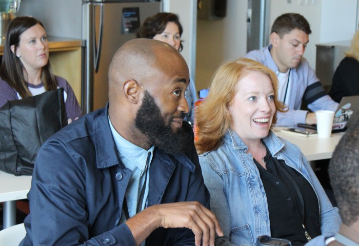 An African American man with a beard and a caucasian woman with red hair sit in a crowd. They are listening for more information.